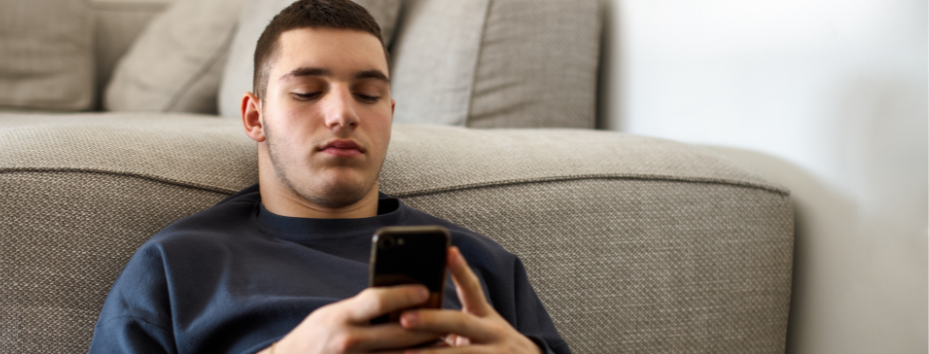 young man looks at phone, lying on the floor propped against sofa
